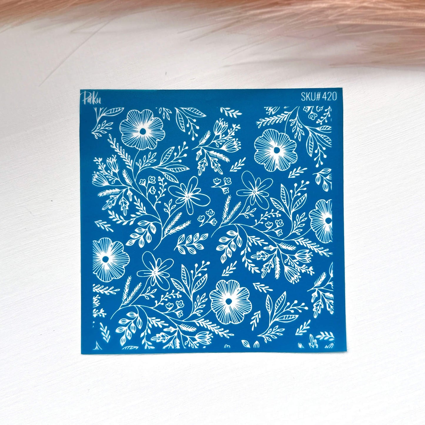 A square blue stamp or silkscreen stencil with a white floral and leaf pattern, placed on a light surface with a soft pink feather in the background.