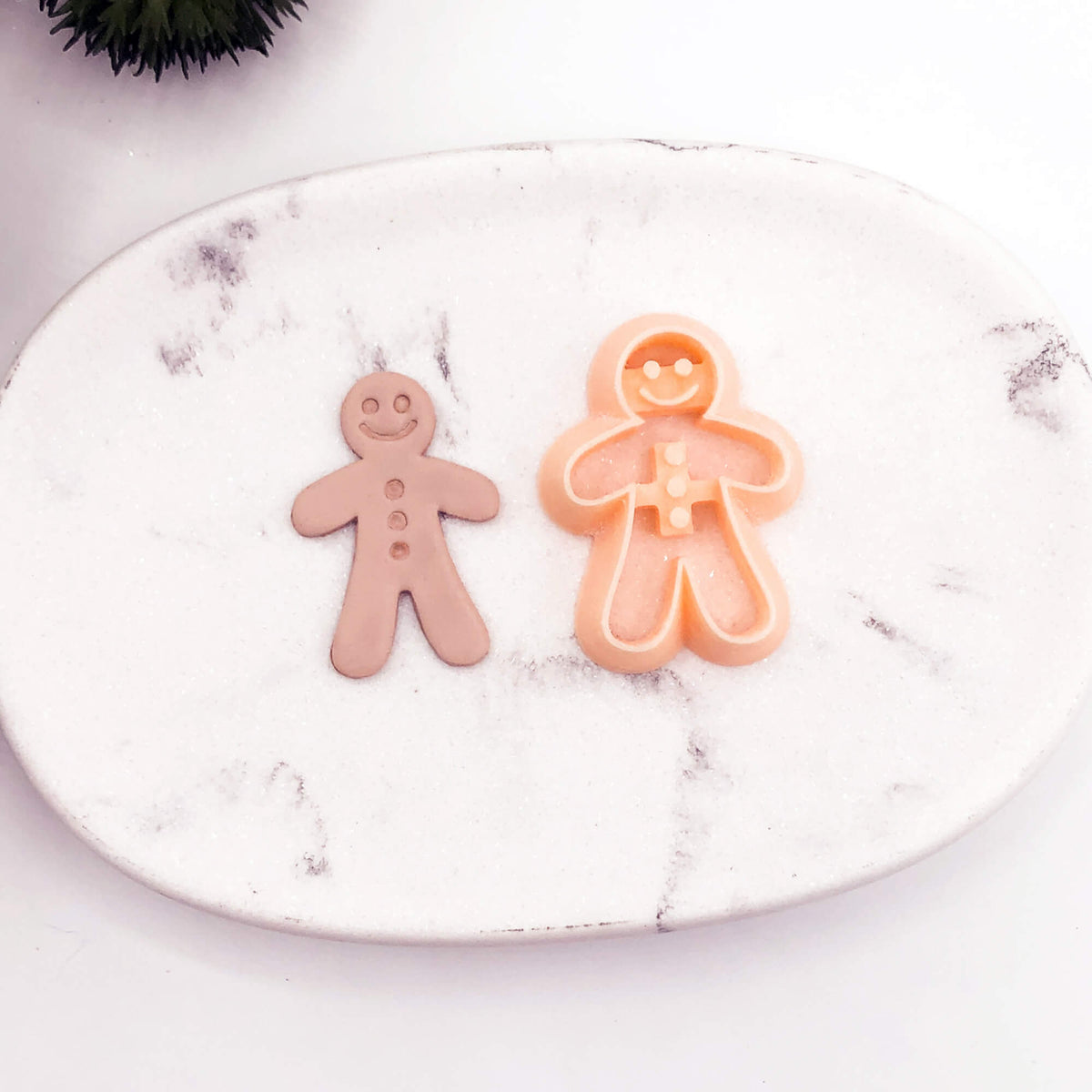 A gingerbread man-shaped clay cookie cutter with a clay cut out displayed on a white marble tray.