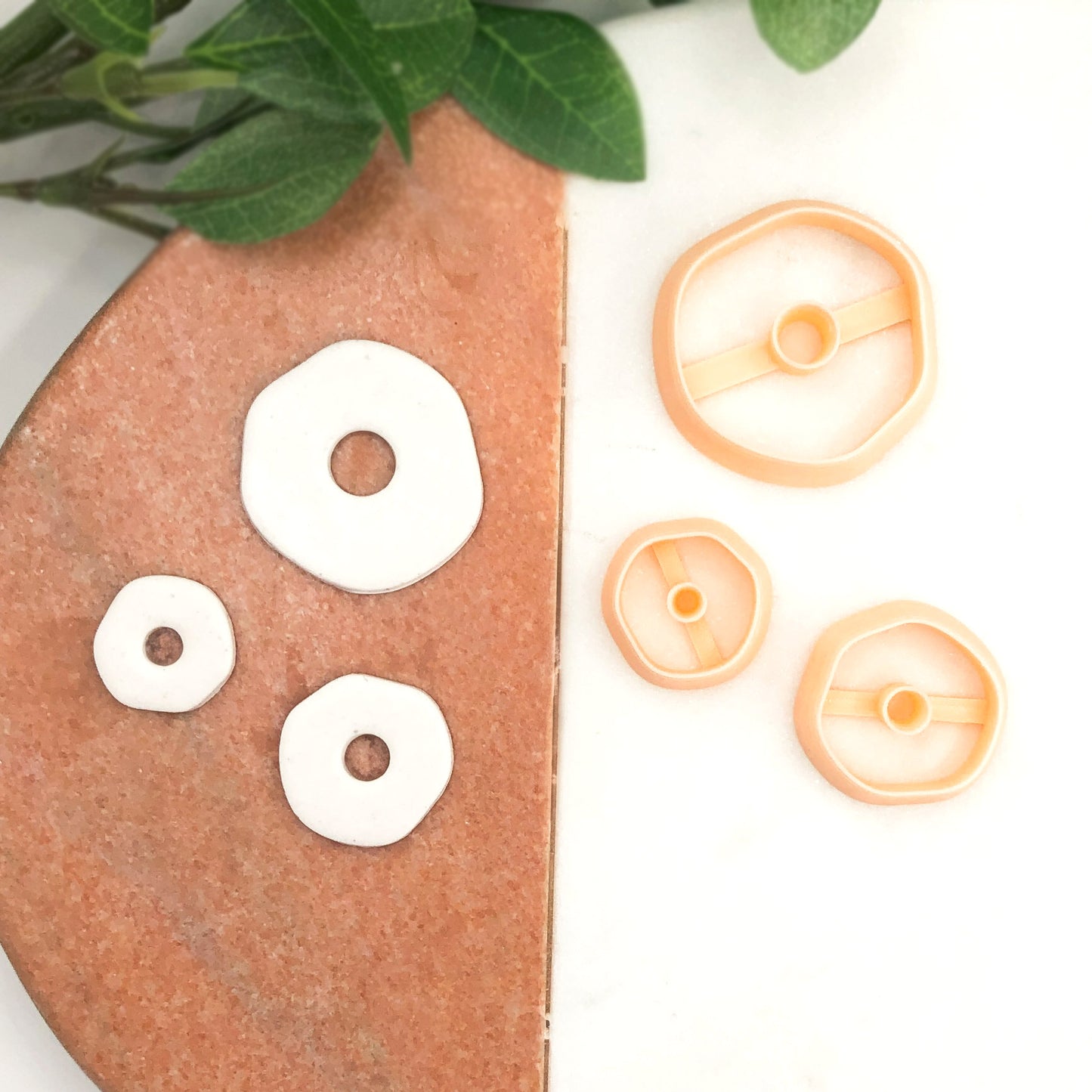 Three peach-colored plastic clay cookie cutters in different sizes are displayed on a white surface, with three of them shown pressing into white clay on a terracotta-colored stone slab and green leaves in the top left corner.