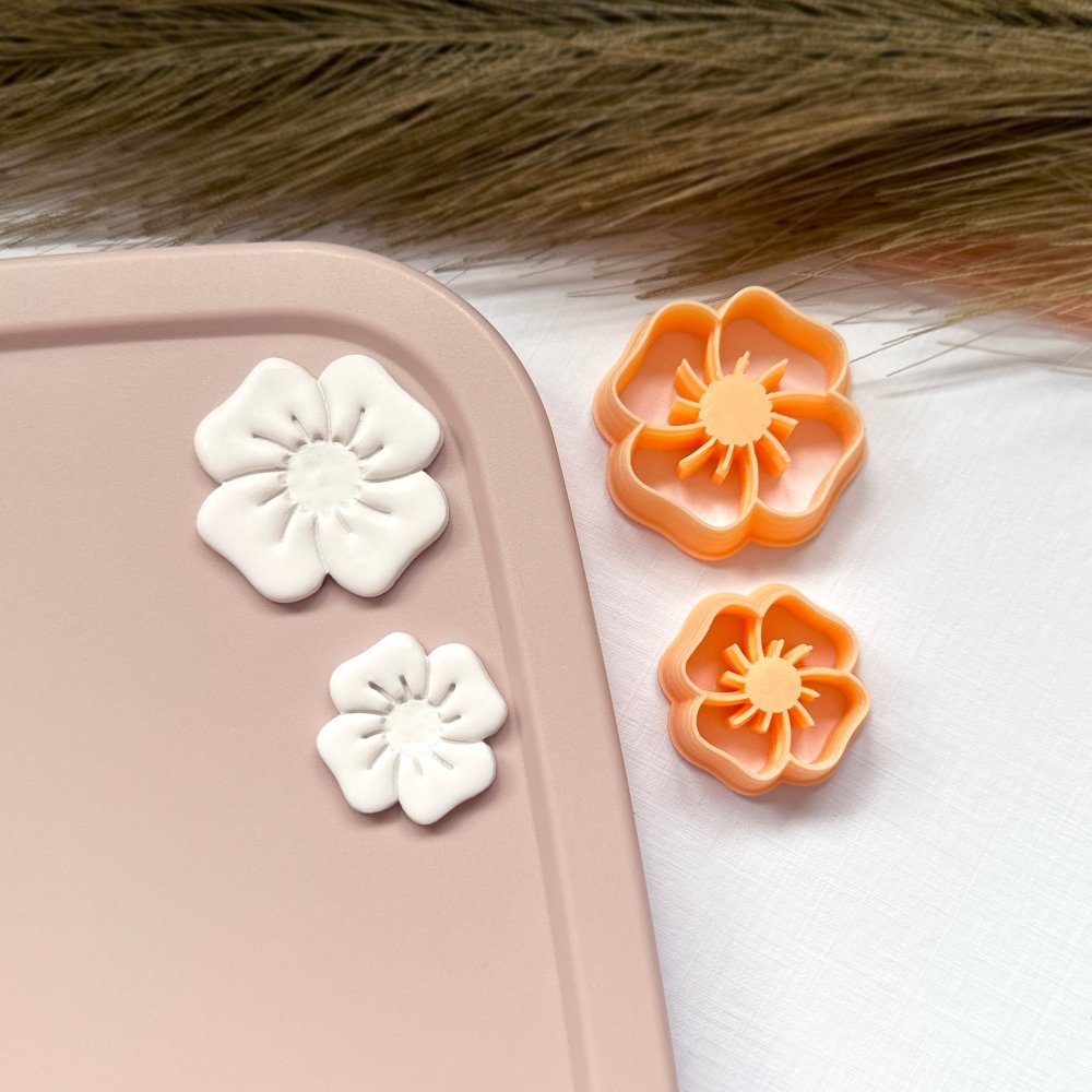 Poppy Clay Cutter - white polymer clay flower poppies shown on a hand, next to the orange poppy-shaped clay cutters they were made with.