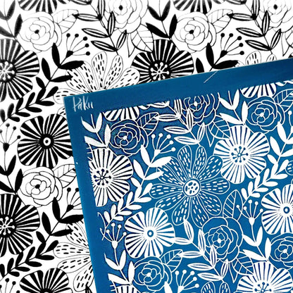 A square blue card or silkscreen stencil with a white floral line-art pattern, placed over a black-and-white floral patterned background. The blue card has a small visible brand mark reading 'Hikaru' in the upper left corner.