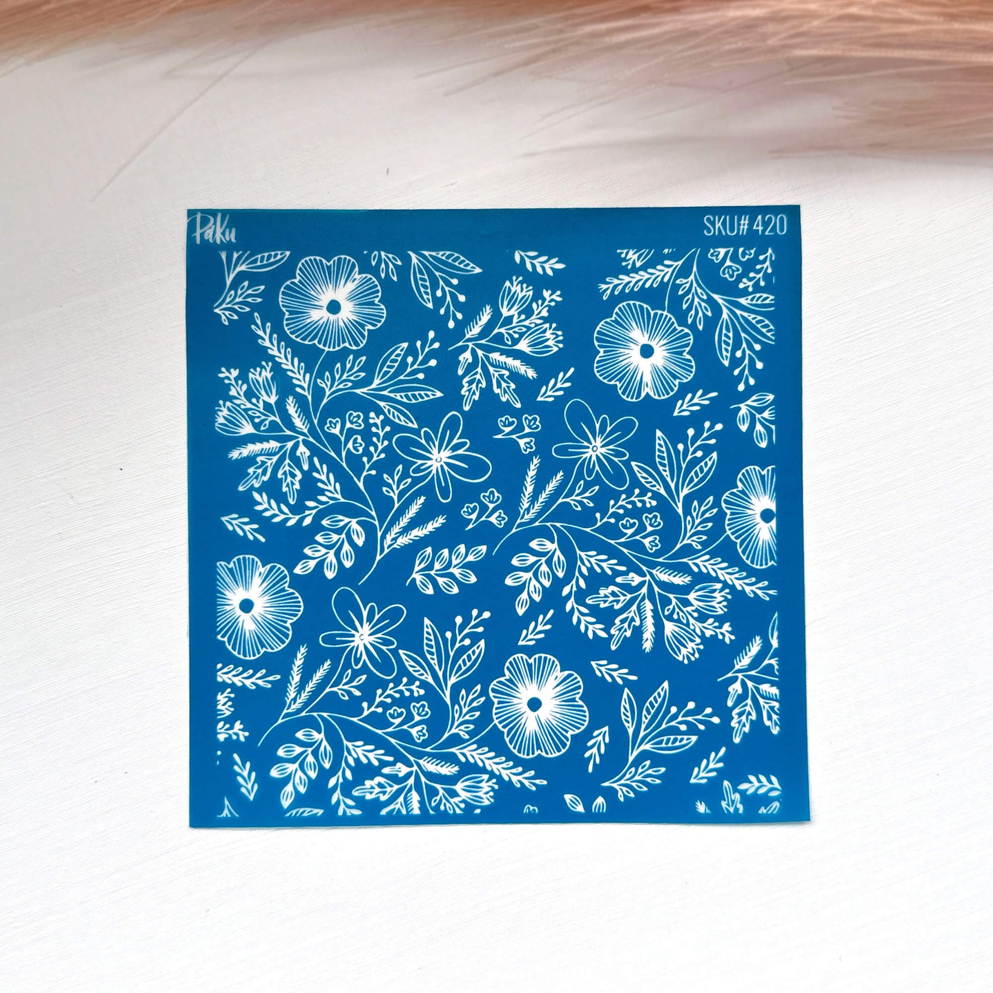 A square blue stamp or silkscreen stencil with a white floral and leaf pattern, placed on a light surface with a soft pink feather in the background.