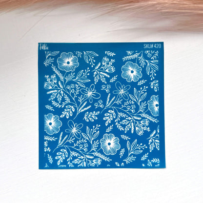 A square blue stamp or silkscreen stencil with a white floral and leaf pattern, placed on a light surface with a soft pink feather in the background.