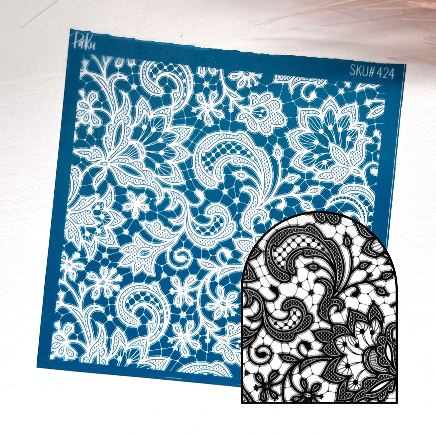 A blue rectangular lace silkscreen stencil template with an intricate white floral and paisley cutout pattern, shown on a light wood background. A smaller inset at the bottom right shows the black-and-white lace design created by cutting through the stencil.
