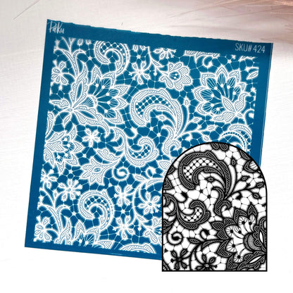A blue rectangular lace silkscreen stencil template with an intricate white floral and paisley cutout pattern, shown on a light wood background. A smaller inset at the bottom right shows the black-and-white lace design created by cutting through the stencil.