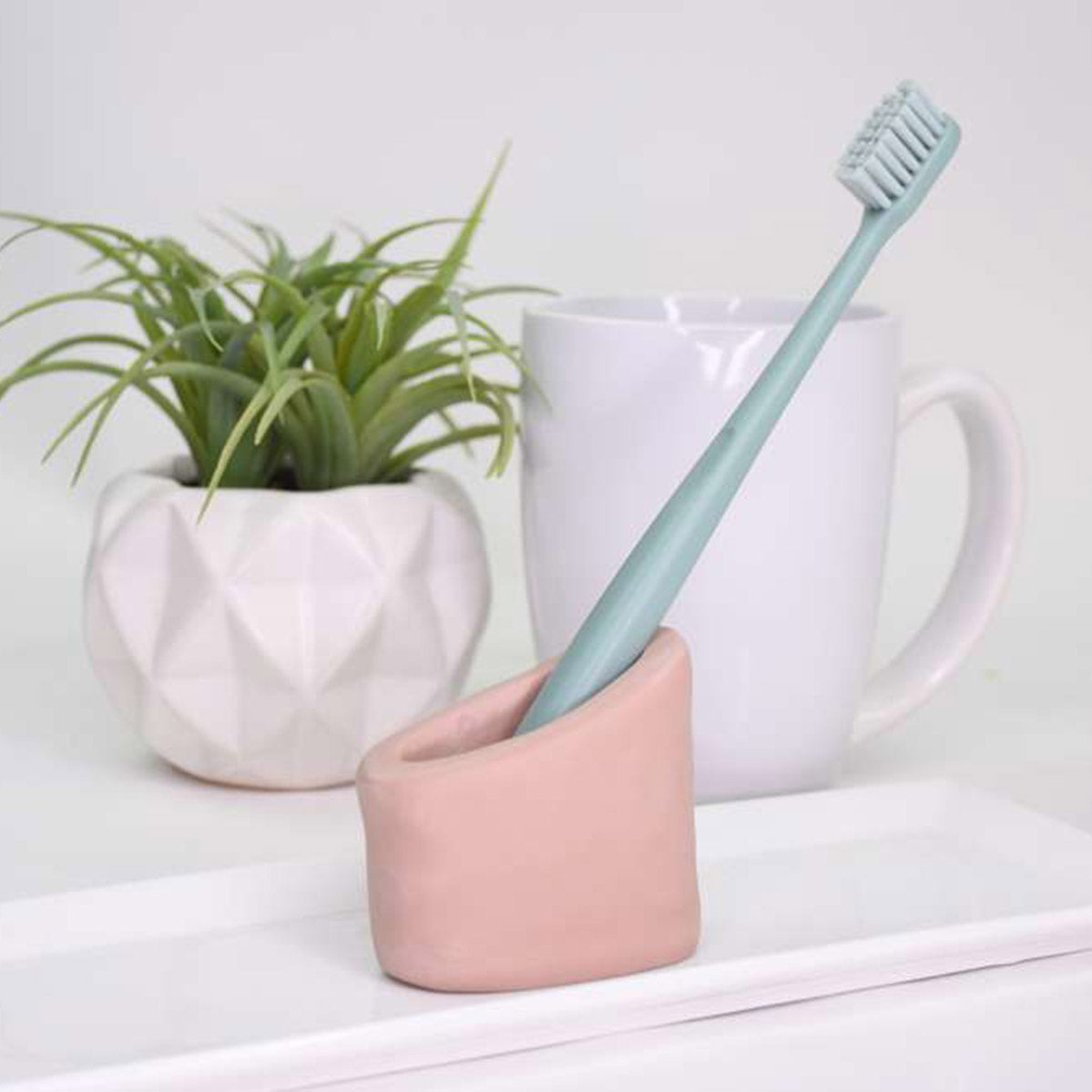 Light blue toothbrush in a pink holder on a white surface with a plant and white mug in the background. made with Sculpey Souffle Polymer Clay | Amond Rose - 48g - 