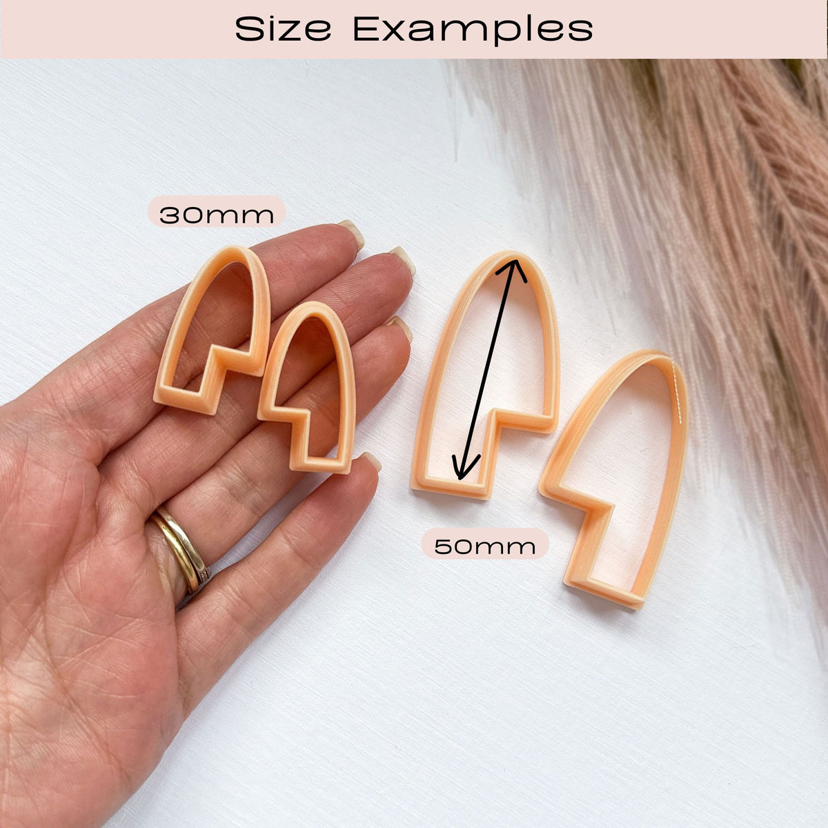 A hand holds two peach-colored arch-shaped cookie clay cutters labeled 30mm, with two larger matching cutters shown on a white surface labeled 50mm. The image includes a header reading 'Size Examples' and a soft pink grass prop on the right.