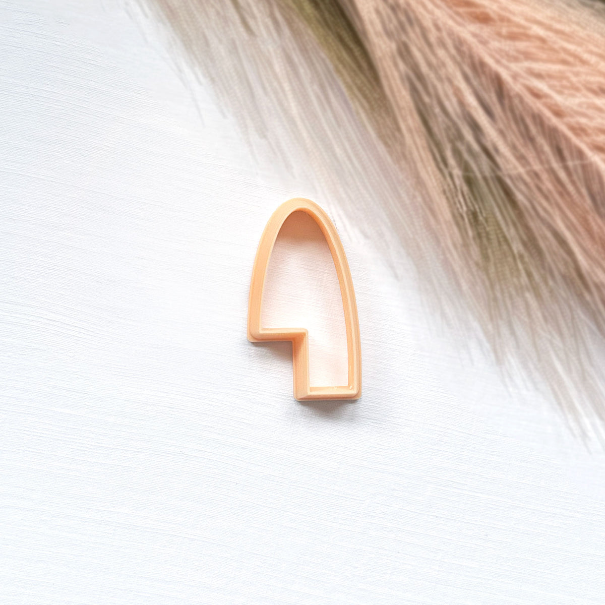 A small peach-colored arch-shaped cookie clay cutter placed on a white textured surface with dried grass in the upper right corner.