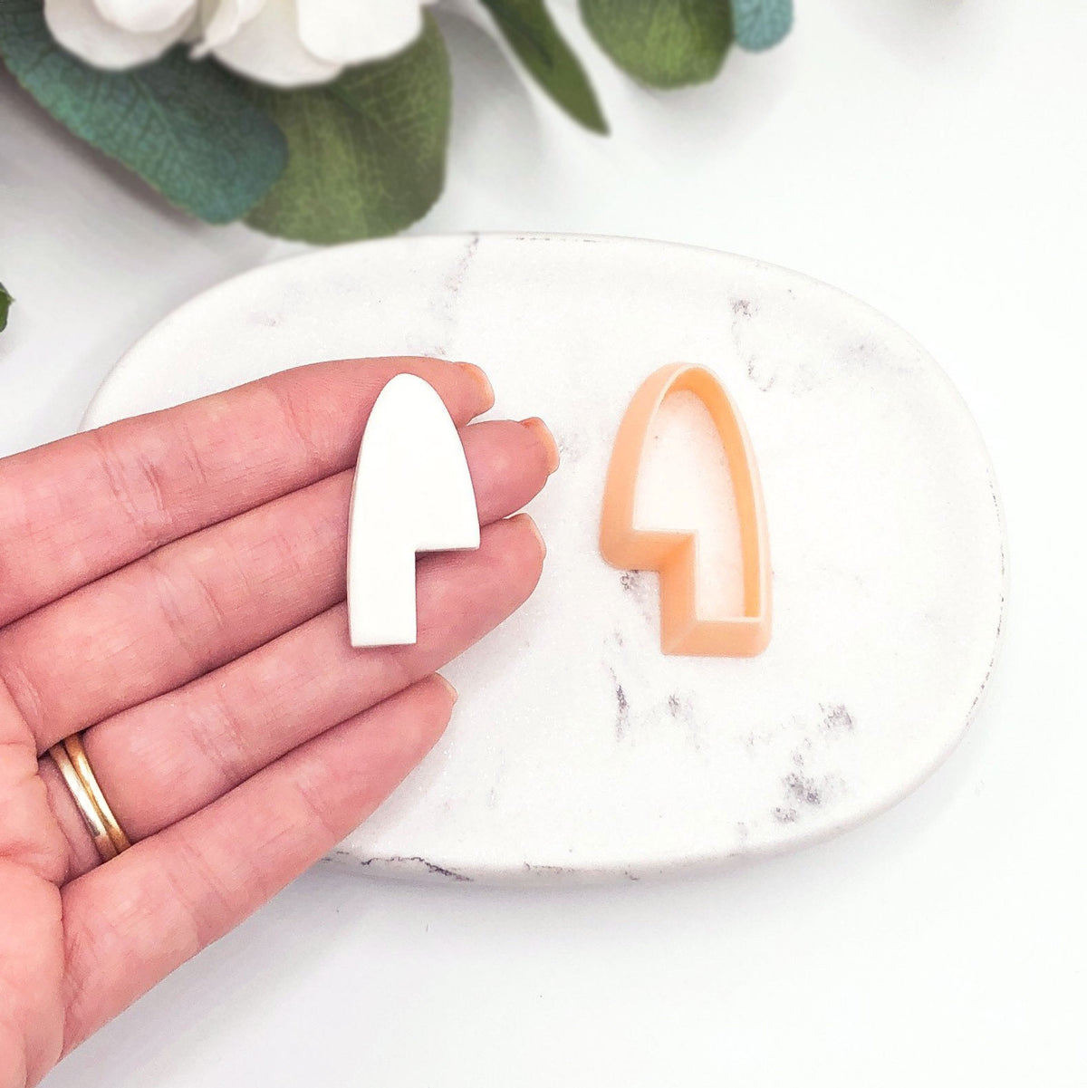 A hand holds a small white arch-shaped item next to a peach-colored arch cookie clay cutter on a white marble dish, with blurred green leaves in the background.