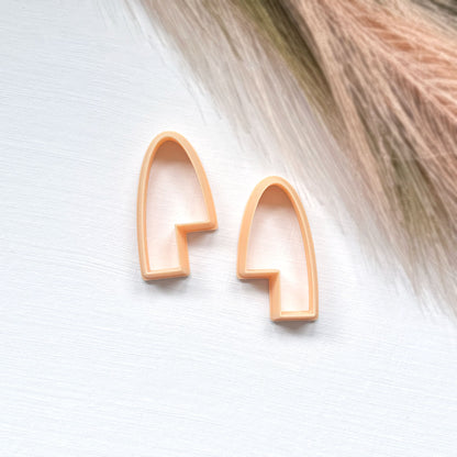 A pair of mirrored arch-shaped clay cutters in a warm peachy-orange color, displayed on a light textured white surface with a soft beige and brown grass prop in the upper right corner.