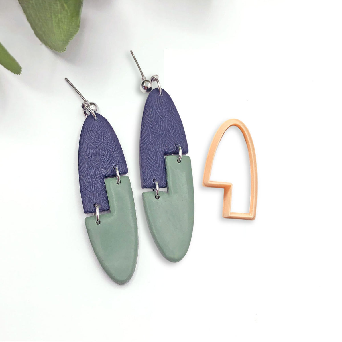 A pair of oval dangle earrings with a geometric interlocking design. The earrings are made of two arch stepped shape pieces of polymer clay, one dark blue with a textured pattern and one light green. The earrings are attached to silver ear wires. The single clay cutter used to make the shapes is sat next to the earrings.
