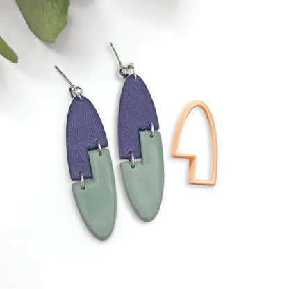 A pair of oval dangle earrings with a geometric interlocking design. The earrings are made of two arch stepped shape pieces of polymer clay, one dark blue with a textured pattern and one light green. The earrings are attached to silver ear wires. The single clay cutter used to make the shapes is sat next to the earrings.