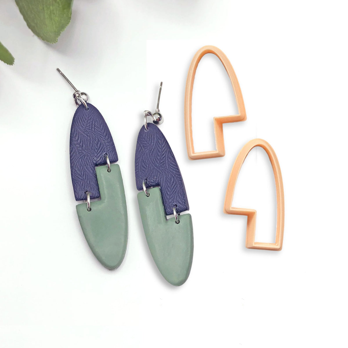 A pair of oval dangle earrings with a geometric interlocking design. The earrings are mirrored and made of two arch stepped shape pieces of polymer clay, one dark blue with a textured pattern and one light green. The earrings are attached to silver ear wires. The mirror set of clay cutters used to make the shapes are sat next to the earrings.