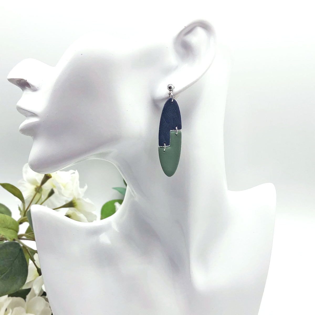 A pair of long oval drop earrings with a dark navy blue top half and a light green bottom half, separated by small silver loops, displayed on a white female mannequin head.