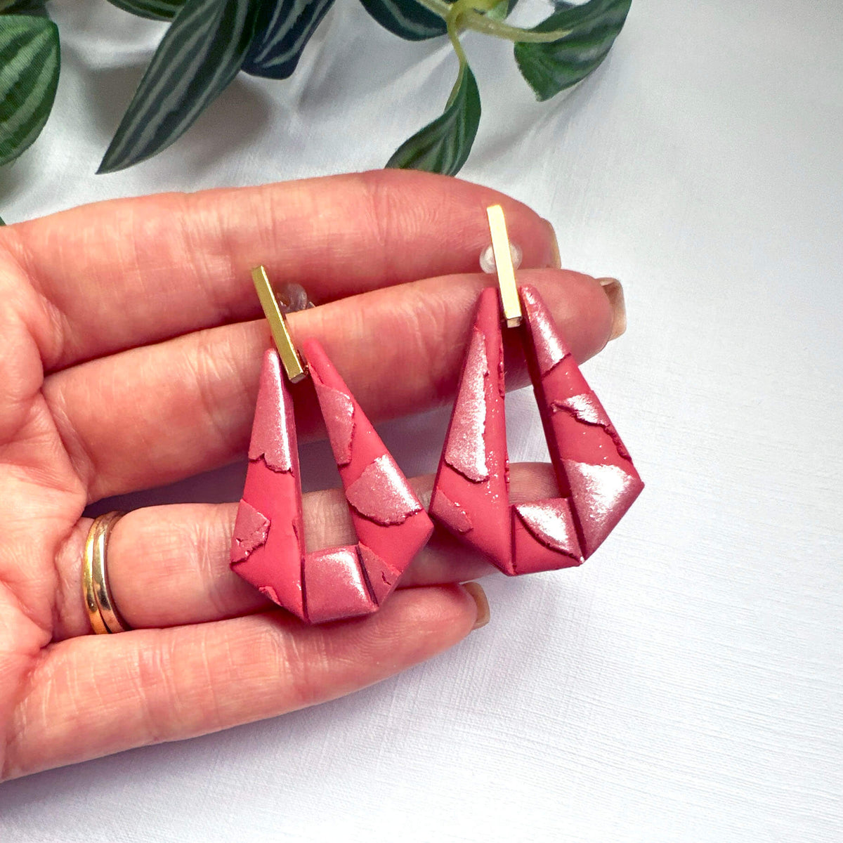 A pair of handmade geometric drop earrings held in a person's hand. The earrings feature a textured, glossy pink-red triangular design with gold-tone posts, shown against a white background with green leaves in the upper corner.