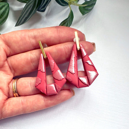 A pair of handmade geometric drop earrings held in a person's hand. The earrings feature a textured, glossy pink-red triangular design with gold-tone posts, shown against a white background with green leaves in the upper corner.