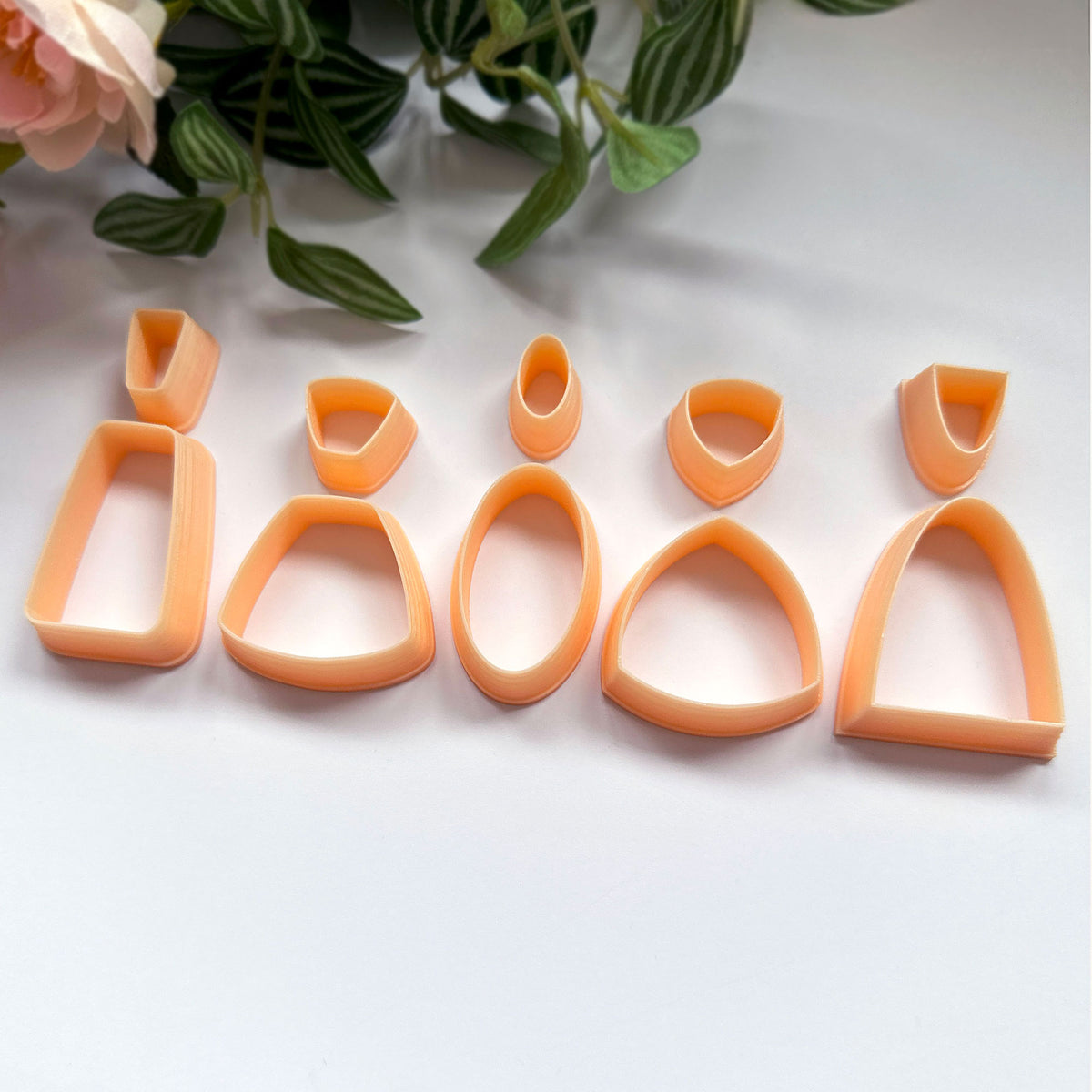 A set of 10 peach-colored 3d printed cookie clay cutters in various shapes, arranged on a white surface with decorative green leaves and a pink rose in the background.