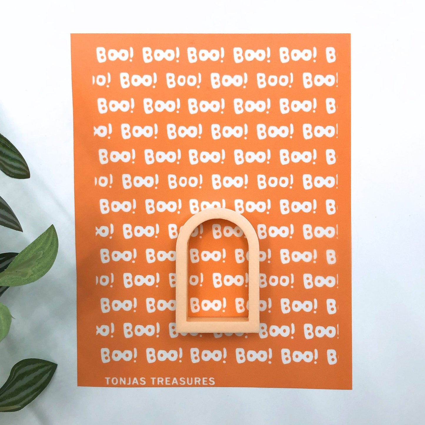 An orange Halloween-themed silkscreen with repeated white 'BOO!' text and a wooden arch-shaped ring placed on top, photographed against a white background with a leaf at the left edge. The brand name 'TONJAS TREASURES' appears at the bottom.