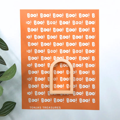 An orange Halloween-themed silkscreen with repeated white 'BOO!' text and a wooden arch-shaped ring placed on top, photographed against a white background with a leaf at the left edge. The brand name 'TONJAS TREASURES' appears at the bottom.