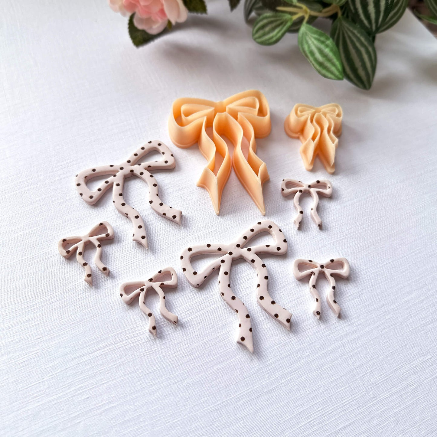 Bow-shaped polymer clay cut-outs with polka dots on a white surface with green leaves in the background and the clay cutters 