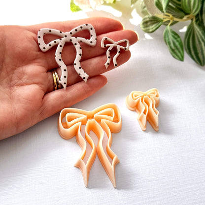 Hand holding bow-shaped polymer clay cut-outs on a light surface with the clay cutters in the front and a plant in the background
