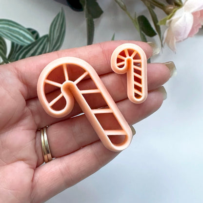 A hand holding two peach-colored plastic candy cane-shaped cookie cutters with a soft-focus background of green leaves and pale pink flowers.