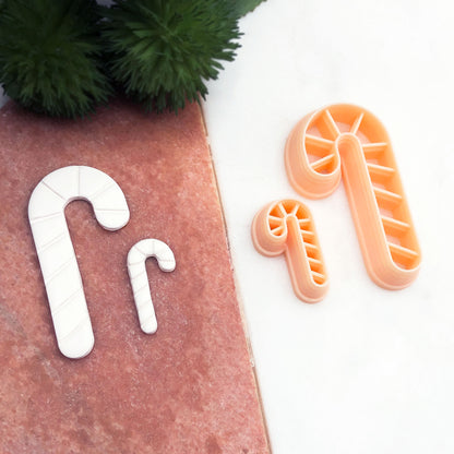Two candy cane-shaped cookie cutters, in peach, displayed on a split white and terracotta background with white clay cut outs of the matching shape and green pine branches in the corner.