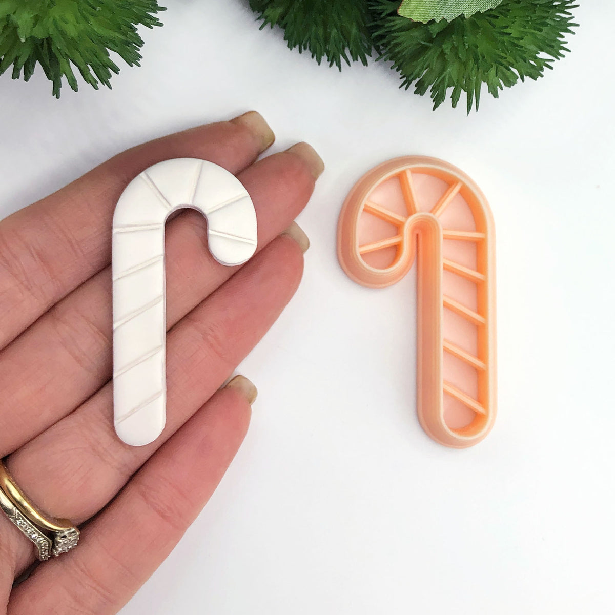 A hand holds a white candy cane-shaped clay cut out next to an orange plastic cookie cutter shaped like a candy cane on a white background with green foliage at the top.