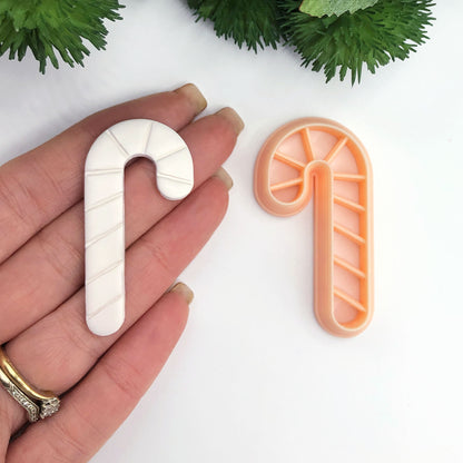 A hand holds a white candy cane-shaped clay cut out next to an orange plastic cookie cutter shaped like a candy cane on a white background with green foliage at the top.