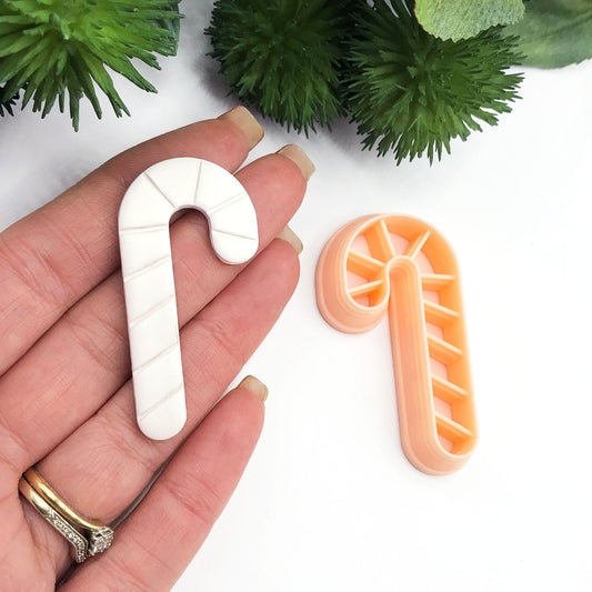 A hand holds a white candy cane-shaped clay cut out while a peach-colored matching cutter lies beside it on a white surface with green decorative foliage in the background.