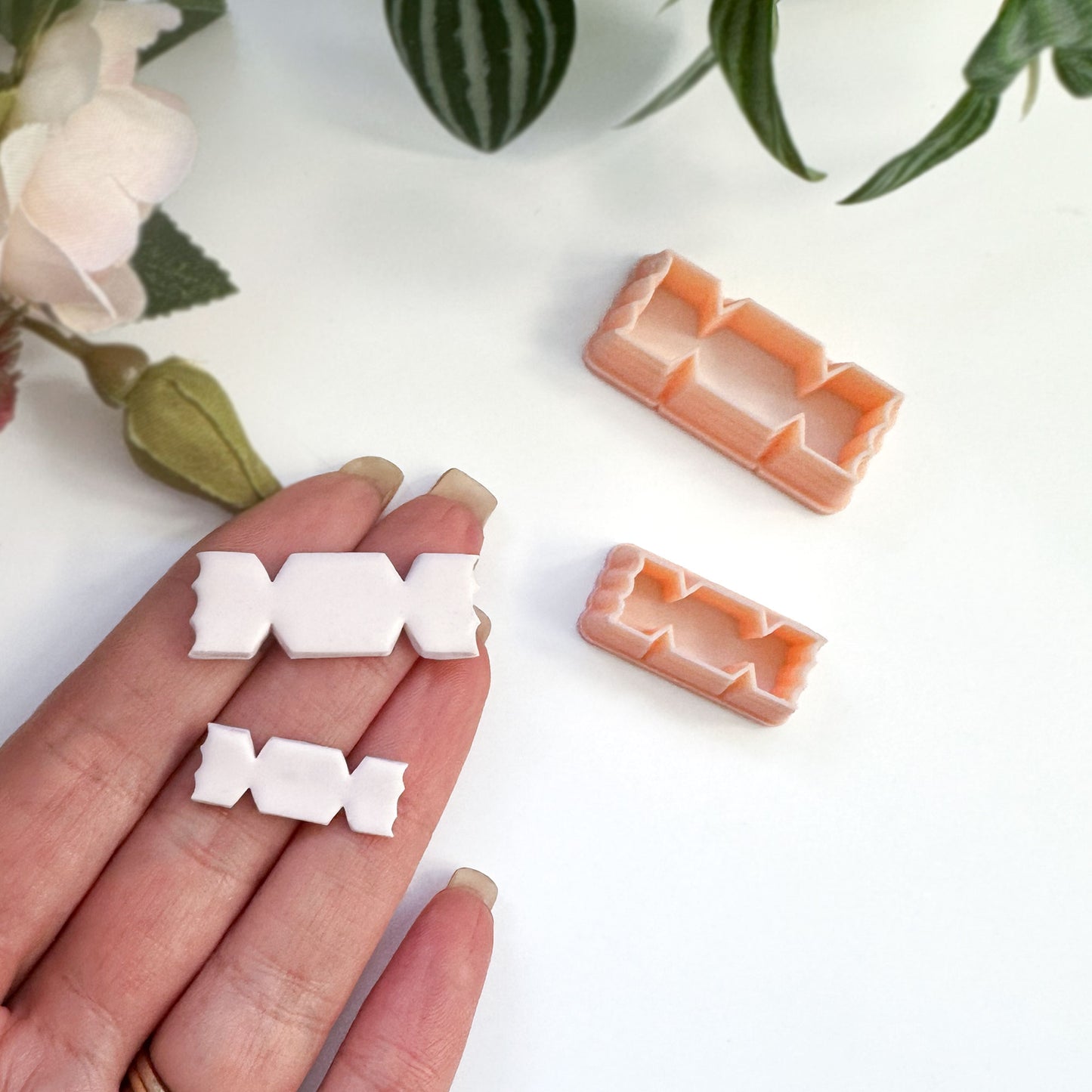 A hand holds a small white candy-shaped cut out in polymer clay, with an orange plastic matching cutter placed nearby on a light background.