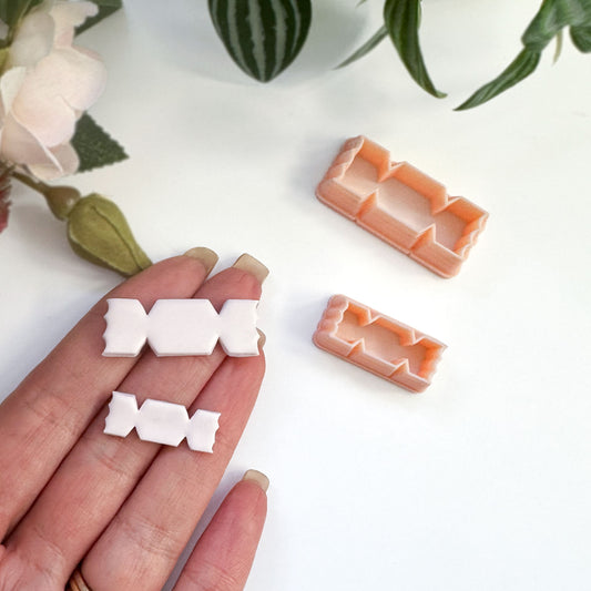 A hand holds a small white candy-shaped cut out in polymer clay, with an orange plastic matching cutter placed nearby on a light background.