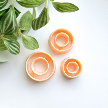 Three peach-colored plastic ring-shaped clay cutters arranged on a white surface with green plant leaves in the upper left corner.