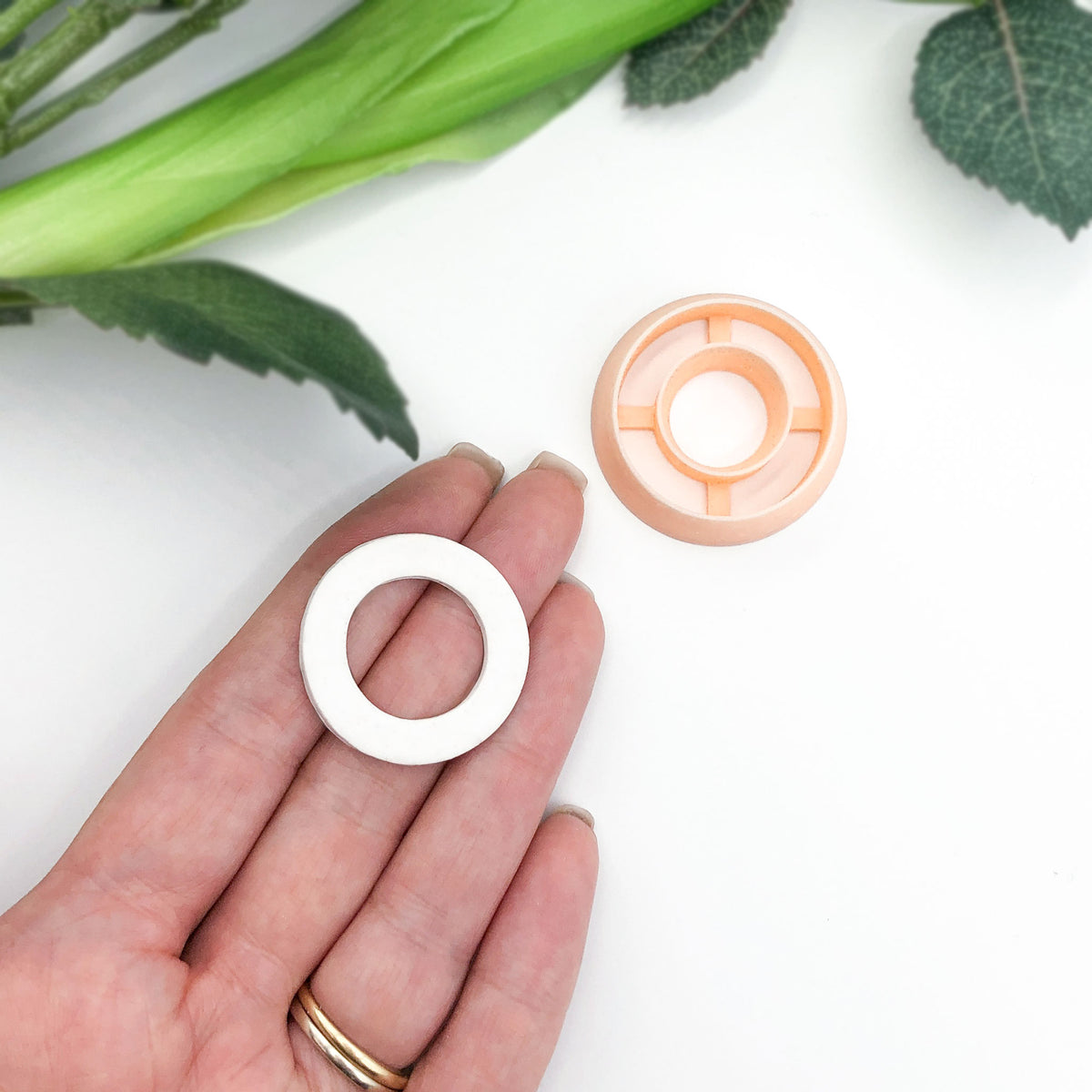 A hand holds a small white circular ring-shaped clay cut out next to a matching peach-colored plastic clay cutter on a white background with green leaves in the corner.
