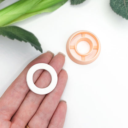A hand holds a small white circular ring-shaped clay cut out next to a peach-colored plastic clay cutter on a clean white surface with green leaves in the background.