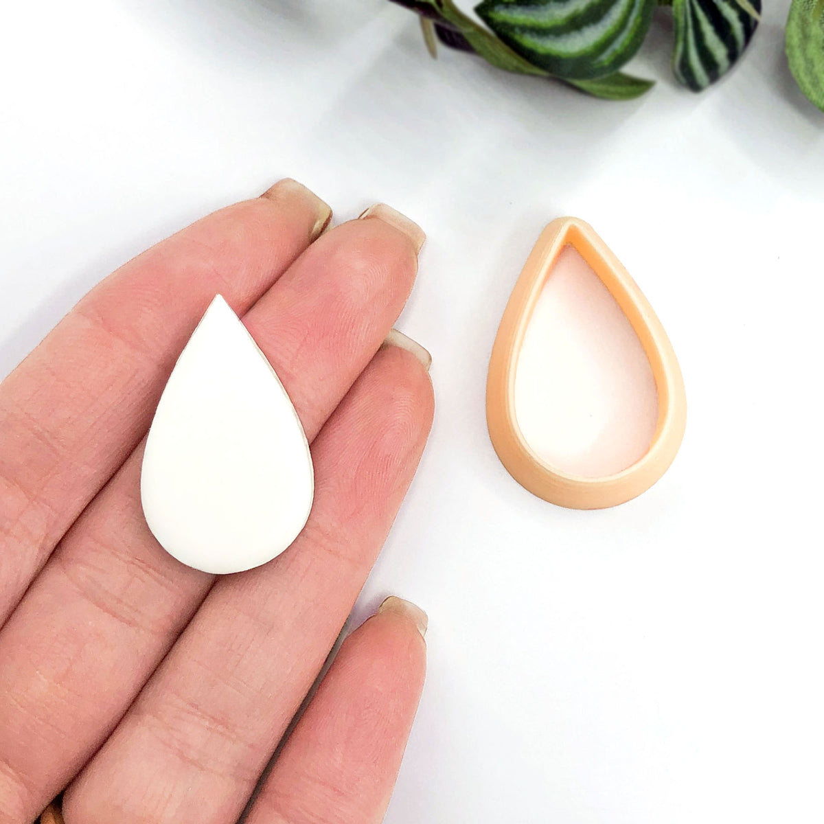 A white teardrop-shaped polymer clay cut out shape and a matching peach clay cutter. Shaped like a petal or raindrop.