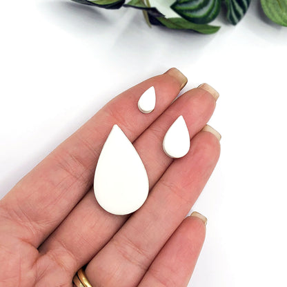 Three white teardrop-shaped clay blanks in different sizes: 10mm, 15mm, and 30mm. The blanks are held in a hand for size comparison.