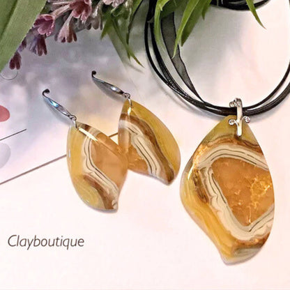 A jewelry set featuring a teardrop-shaped pendant necklace and matching drop earrings made from polymer clay faux marbled agate with warm yellow, orange, and cream banding, displayed on a white surface with green leaves and flowers in the background. The text 'Clayboutique' is visible in the lower left.