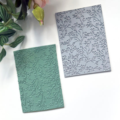 A rectangular embossed rubber stamp with a delicate coral leaf pattern, shown in sage green and light gray on a white background with decorative green leaves and a pink flower in the corner.