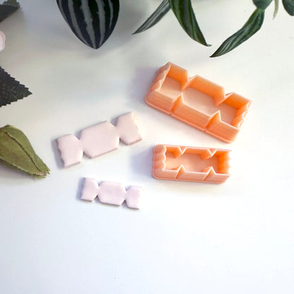 Two peach-colored plastic cookie cutters in a sweetie designs, shown alongside three pale pink cutout pieces on a white surface with decorative green leaves.