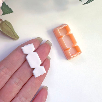 A hand holds a small white candy-shaped cut out in polymer clay, with an orange plastic matching cutter placed nearby on a light background.