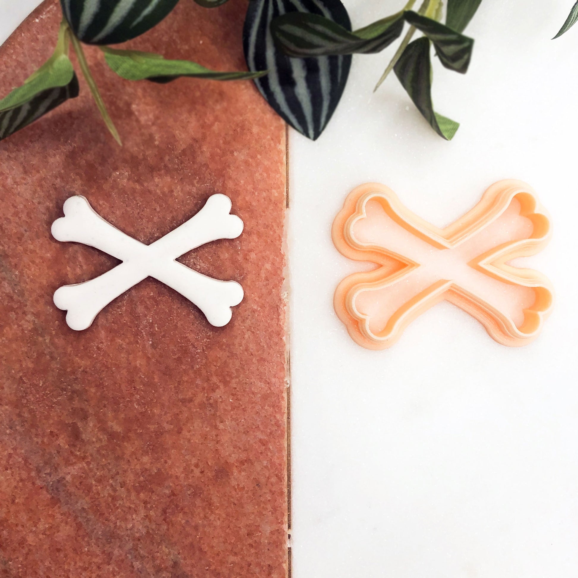 A peach plastic cookie clay cutter in the shape of crossed bones. The cutter is shown next to a white clay piece that has been cut out using the cutter.