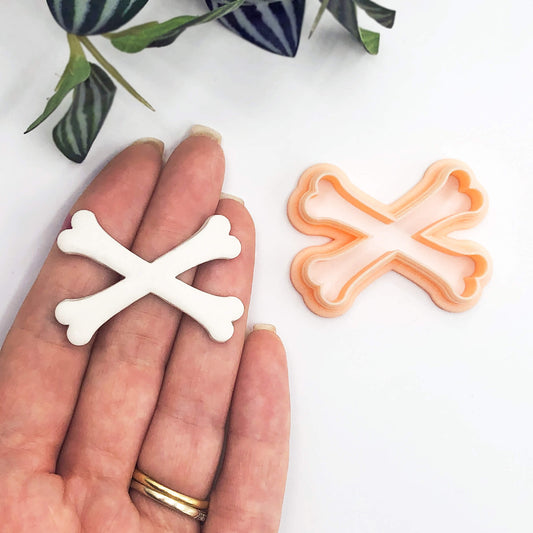 A peach plastic cookie clay cutter in the shape of crossed bones. The cutter is shown next to a white clay piece that has been cut out using the cutter.