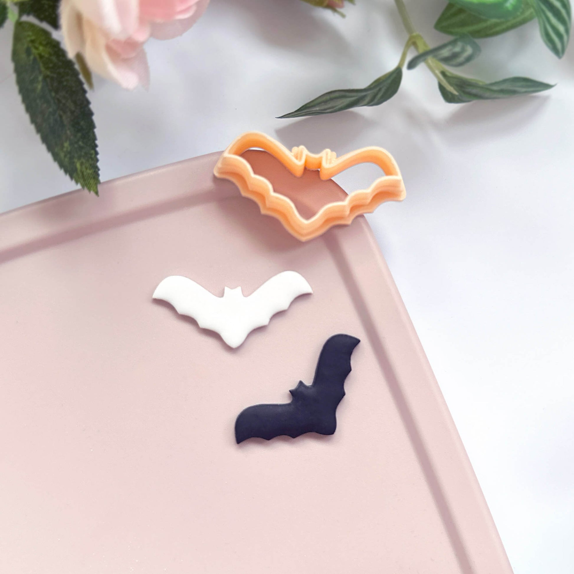 Three bat-shaped cookie cutters in white, dark navy, and peach-orange are displayed on a light pink tray with decorative flowers in the corner.