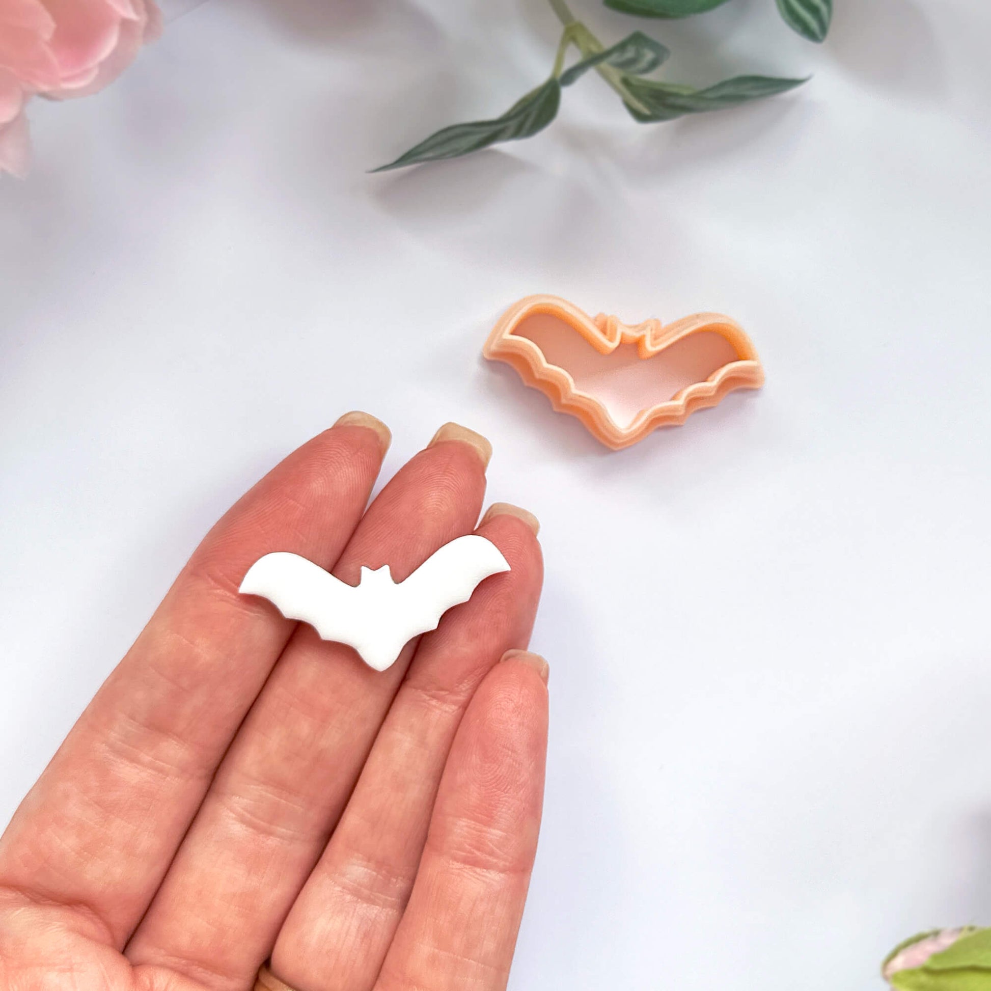 A hand holds a small white bat-shaped polymer clay cut out, with an orange bat-shaped clay cookie cutter placed nearby on a light background. Pink flowers and green leaves are softly blurred in the corners.