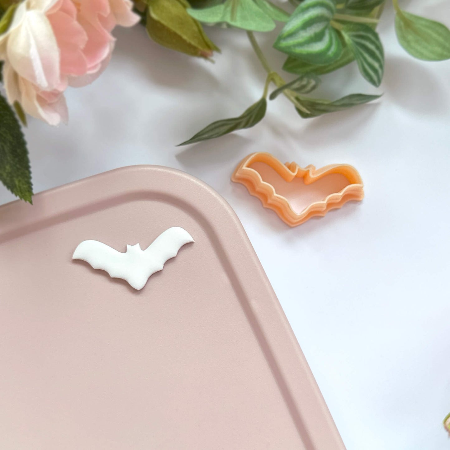 A small bat-shaped clay cookie cutter in orange and a white bat-shaped piece of polymer clay, placed on a pale pink tray with decorative pink flowers and green leaves in the background.