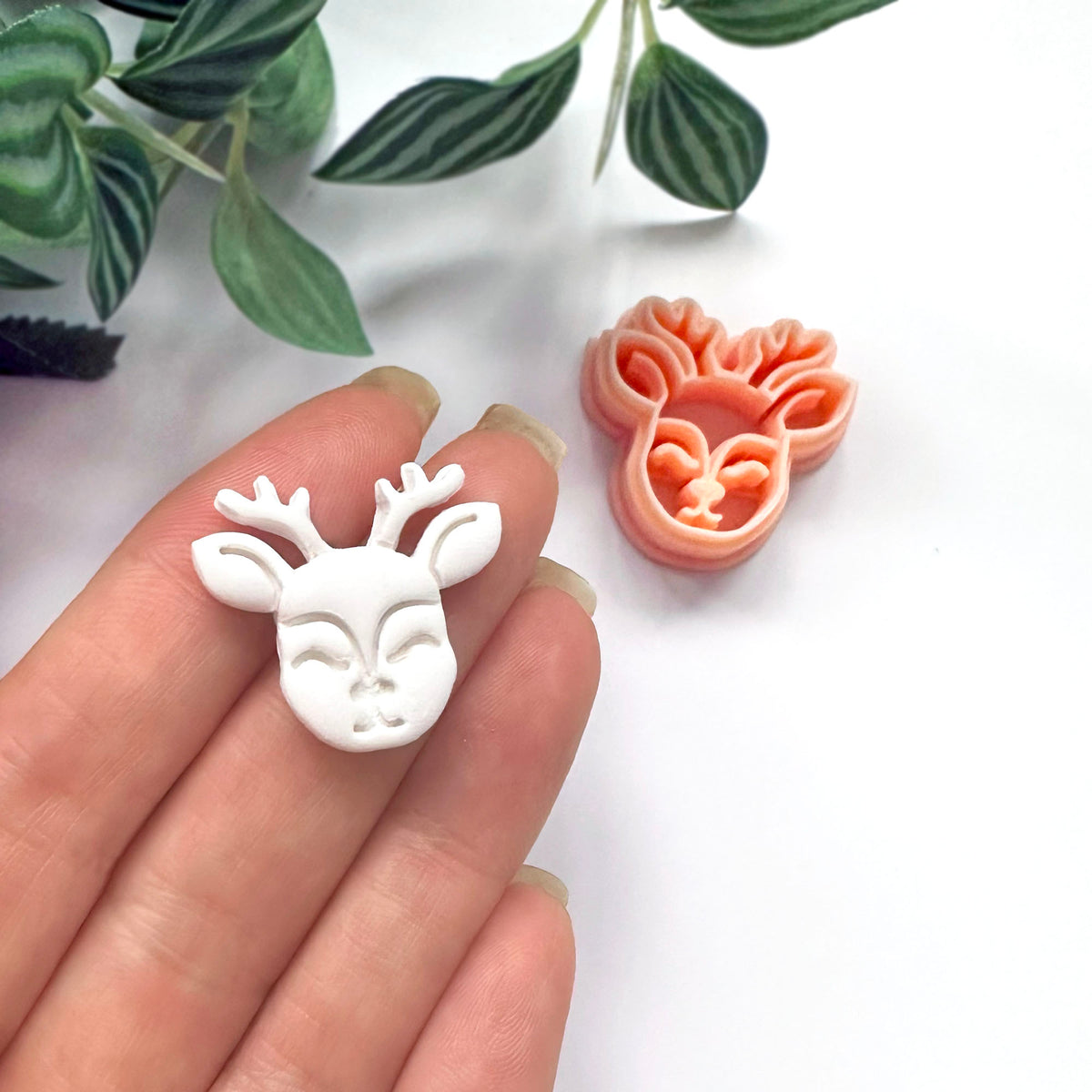 A hand holds a small white deer head polymer clay charm while a peach-colored matching clay cutter sits on a white surface, with green leaves in the background.