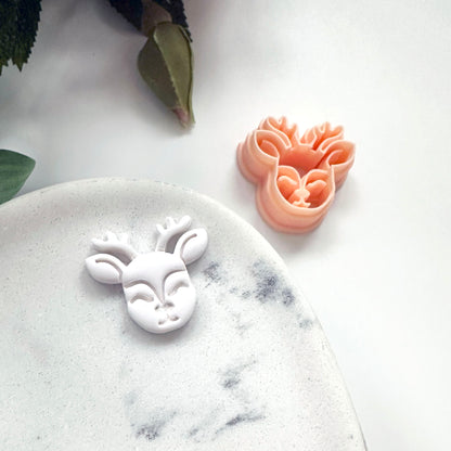 A small deer head cookie cutter in peach pink and a matching white cutout sample on a white marble dish, with a white background and a leaf in the corner.