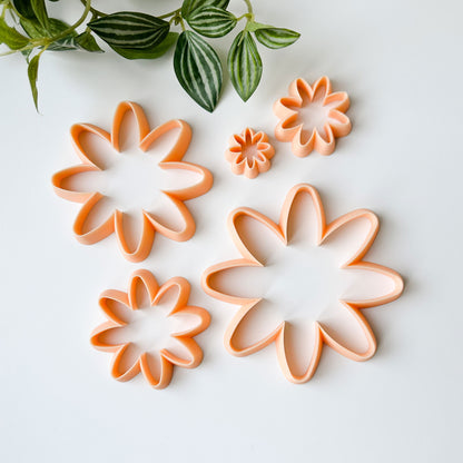 Four daisy flower-shaped cookie cutters in a peach color arranged on a white background, with a green leafy plant visible in the top left corner.