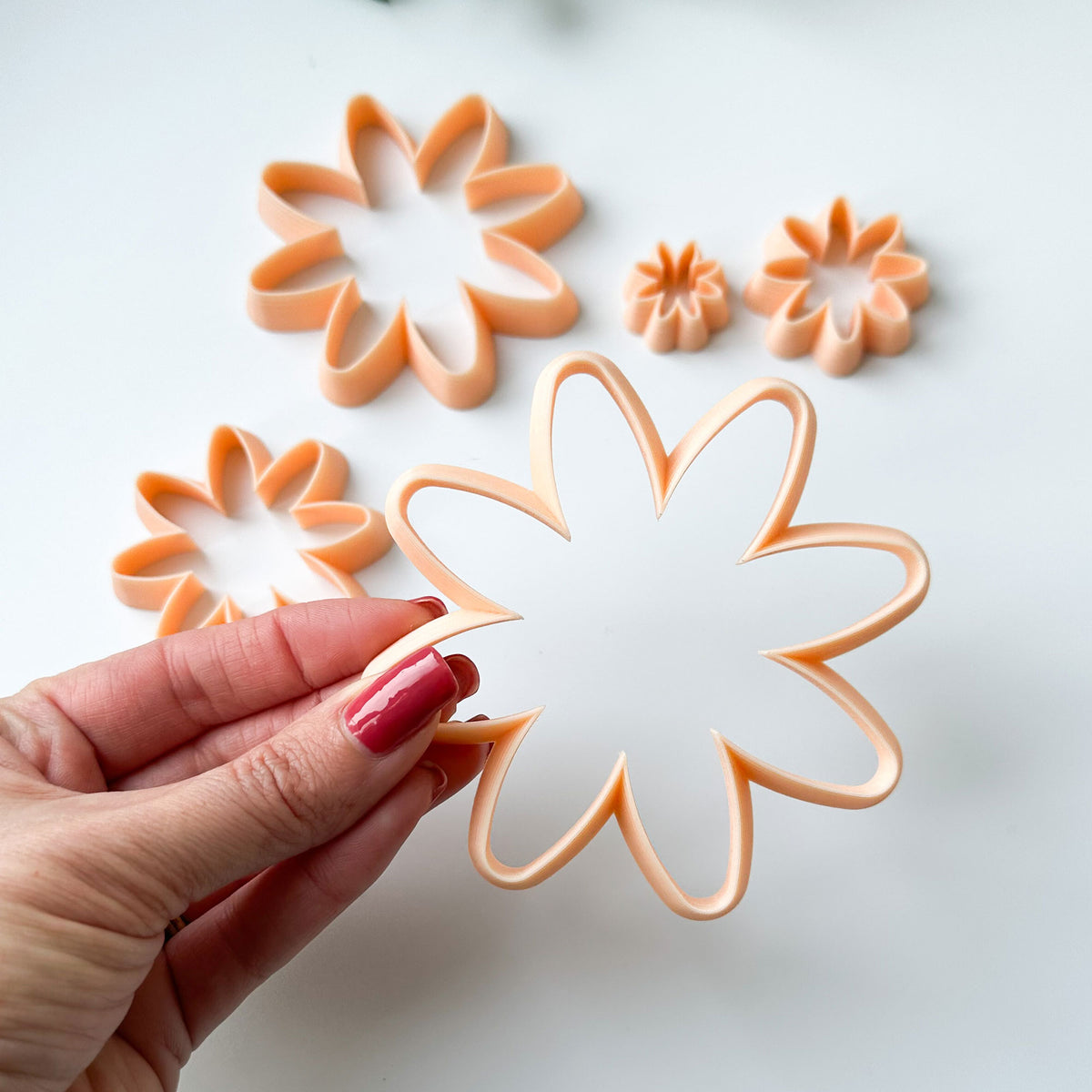 A hand with red nail polish holds a peach-colored plastic flower-shaped cookie cutter, with four larger matching flower cutters arranged on a clean white background.